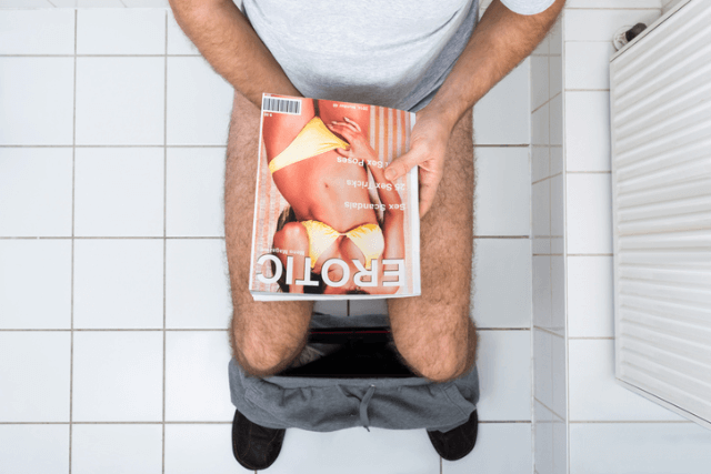 ズボンを脱ぎ雑誌で陰部を隠してトイレに座っている男性